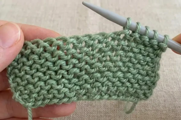 Imagem de uma peça de tricô em andamento, feita com fio verde claro, envolvendo agulhas de tricô, mostrando também pontos de trico